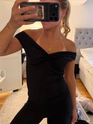 Svart offshoulder topp - Säljer en svart offshoulder topp med twistad detalj framtill. Toppen är tight och har en smickrande passform som framhäver axlarna. Perfekt att styla med både jeans och kjol för en trendig look.