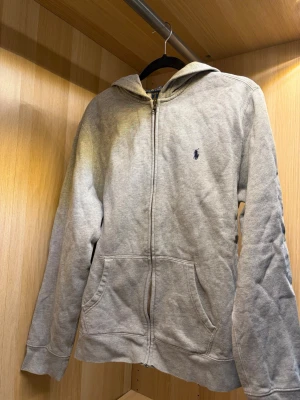 Grå zip hoodie från polo Ralph lauren - En stilren grå hoodie med dragkedja och huva. Klassisk passform med två stora fickor framtill och diskret logga broderad på bröstet. Perfekt för chill dagar eller när du vill ha en enkel och snygg look. Mjuk bomullskänsla och ribbade muddar vid ärmar och nederkant.
