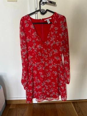 Röd blommig kortklänning NLY Trend - Snygg röd kortklänning från NLY Trend med blommigt mönster i vitt och grått. Klänningen har v-ringning, långa ärmar och dragkedja i ryggen. Perfekt för sommarens alla tillfällen!