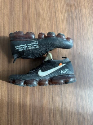 Nike Off-White Air Vapormax svart - Säljer ett par Nike Off-White Air Vapormax i svart med vit Swoosh och textdetaljer på sidan. Skorna har genomskinlig bubbelsula, mesh- och textilmaterial samt orange Off-White tag. Riktigt ikonisk och streetinspirerad sneaker.