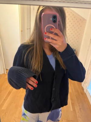 Mörkblå stickad kofta med knappar - Mysig mörkblå stickad kofta med v-ringning och stora knappar framtill. Koftan har långa ärmar med breda ribbade muddar och en avslappnad passform. Perfekt att slänga över en t-shirt för en chill look.