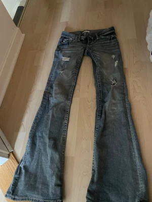 Ljusblåa slitna bootcut jeans - Säljer ett par ljusblå bootcut jeans med slitningar och hål på benen. Jeansen har låg midja, klassiska fem fickor och snygga detaljer på bakfickorna. Perfekta för dig som gillar en avslappnad och trendig stil.