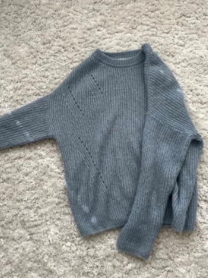 Ljusblå stickad tröja i mohairmix - Mysig ljusblå stickad tröja med rund hals och diskreta hålmönster på framsidan. Tröjan har en fluffig känsla och är troligtvis stickad i en mohair- eller ullmix. Perfekt för kyliga dagar och enkel att styla med jeans eller kjol.