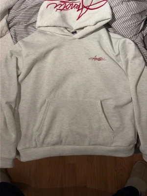 Ljusgrå hoodie med röd logga - Snygg ljusgrå hoodie med röd broderad logga på bröstet och stor röd text på huvan. Klassisk känguruficka framtill och ribbade muddar vid ärmar och nederkant. Perfekt för en avslappnad och trendig stil. Pris kan diskuteras