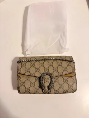  Supreme axelväska - axelväska i GG Supreme-canvas med beige/brunt monogram och mockadetalj framtill. Silverfärgad kedjerem och ikoniskt spänne. Väskan har flera fack och magnetknäppning.