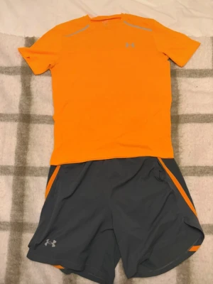 Under Armour träningsset orange/grå - Snyggt träningsset från Under Armour med en kortärmad, neonorange t-shirt och matchande mörkgrå shorts med orange detaljer. Båda plaggen har logga och tryck i reflekterande material. Perfekt för löpning eller gym. Materialet är lätt och andas.