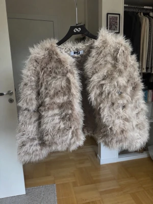 Beige fluffig äkta pälsjacka - Säljer en supermjuk och fluffig beige pälsjacka med lång lugg och rak passform. Jackan har öppen front utan synliga knappar och ger en trendig look. Perfekt för att hålla värmen under kalla dagar och ger en cool statement-stil. Storlek 36/38