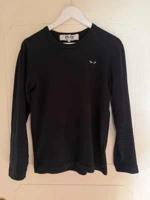 Comme des Garçons långärmad tröja.  - Svart långärmad topp från Comme des Garçons Play med det ikoniska hjärta-ögon-logotypen broderad på bröstet. Tillverkad i mjuk bomull och har en klassisk rund halsringning. Perfekt för en clean och stilren look.
