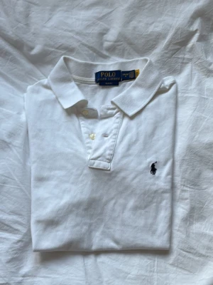 Vit pikétröja från Polo Ralph Lauren - Klassisk vit pikétröja från Polo Ralph Lauren med korta ärmar och krage. Tröjan har knappar framtill och den ikoniska broderade loggan på bröstet. Tillverkad i bomull för en skön och luftig känsla.
