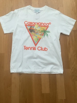 Vit Casablanca Tennis Club t-shirt - Snygg vit t-shirt från Casablanca med färgglatt tryck framtill som visar en retroinspirerad tennisklubb och palmer. T-shirten har rund hals och korta ärmar. Materialet är mjuk bomull och trycket har orange text. Perfekt för en sportig och trendig look.