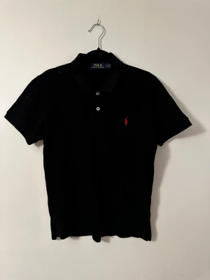 Svart pikétröja från Polo Ralph Lauren - Klassisk svart pikétröja från Polo Ralph Lauren med korta ärmar och två knappar framtill. Tröjan har den ikoniska röda logotypen broderad på bröstet och är tillverkad i mjuk bomull. Perfekt till jeans eller shorts för en clean look. Endast använd fåtal gånger. ( Ny skick i princip) 