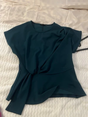Mörkgrön omlottopp med knyt - Stilren mörkgrön topp med omlottdetalj och knyt i sidan. Toppen har korta ärmar och en lös passform som ger en avslappnad men ändå elegant look. Perfekt att matcha med jeans eller kjol för en trendig outfit.