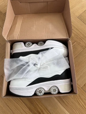 Vita och svarta sneakers med hjul - Unika vita och svarta sneakers med inbyggda hjul i sulan. Skorna har snörning och en sportig design med rund tå och platt sula. Perfekta för dig som vill rulla fram med stil och sticka ut från mängden.