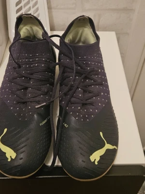 Mörkblåa fotbollsskor från Puma - Säljer ett par mörkblåa fotbollsskor från Puma med stickad ovandel och vita prickdetaljer. Skorna har snörning, strumpliknande skaft och gula Puma-loggor på tårna. Perfekta för dig som vill ha bra grepp och stil på planen.