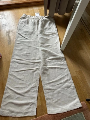 Vida beige linnebyxor med dragsko - Säljer ett par ljusbeige linnebyxor med breda ben och avslappnad passform. Byxorna har resår i midjan och dragsko för extra komfort. Perfekta för en chill och stilren look under varma dagar.
