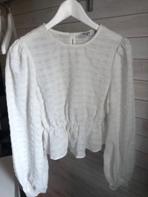 Vit strukturerad blus med volang från NA-KD - Säljer en vit blus med strukturerat tyg och rund hals. Blusen har långa puffärmar och en söt volang nedtill som ger en feminin siluett. Knäppning bak i nacken. Perfekt att styla med jeans eller kjol för en trendig look.