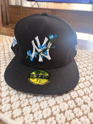 Svart New Era NY keps med blommor - Snygg svart 59FIFTY keps från New Era med New York Yankees-logga framtill, dekorerad med blå broderade blommor. Platt skärm med ljusblå undersida och broderad baseboll. Klassisk snapback-stil och broderade detaljer på sidorna.