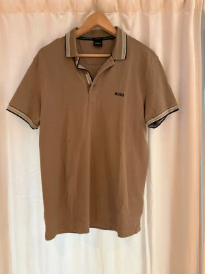 Beige pikétröja från BOSS - Säljer en beige pikétröja från BOSS med klassisk krage och knäppning framtill. Tröjan har korta ärmar och diskreta svarta och vita detaljer på krage och ärmslut. Perfekt för en stilren och avslappnad look.
