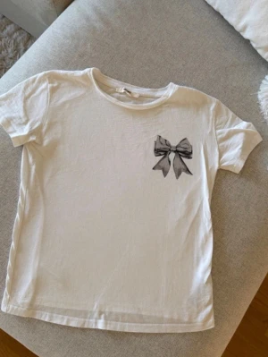 Vit t-shirt med rosettdetalj - Säljer en vit t-shirt med korta ärmar och en snygg tryckt rosett på bröstet. T-shirten har en klassisk rund halsringning och är gjord i mjuk bomull, perfekt för en enkel men söt look.
