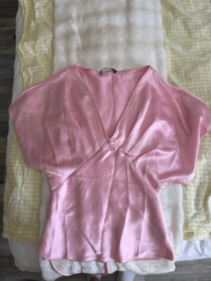 Rosa omlottblus i satin - Snygg rosa omlottblus i glansigt satinmaterial med djup v-ringning och korta, vida ärmar. Figurnära passform i midjan och lösare upptill ger en trendig siluett. Perfekt att styla med jeans eller kjol för en chic look.