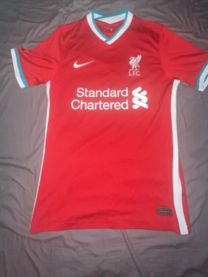 Liverpool FC 2020/21 hemma tröjan - Säljer en röd Liverpool FC matchtröja från Nike med vita och turkosa detaljer vid krage och ärmslut. Tröjan har klubbmärke och sponsortryck på bröstet samt vit sidopanel. Perfekt för dig som älskar fotboll och vill stötta ditt lag.