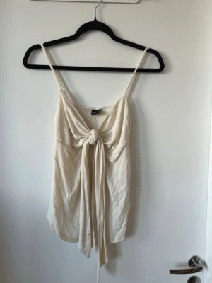 Beige linnetopp med knyt från Gina Tricot - Säljer en beige linnetopp från Gina Tricot med smala axelband och snygg knytning framtill. Toppen är ärmlös och har en avslappnad passform, perfekt för varma dagar. Materialet känns mjukt och lätt mot huden.