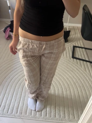Beige rutiga mjukisbyxor med dragsko - Säljer ett par beige mjukisbyxor med vitt rutmönster och dragsko i midjan. Byxorna har avslappnad passform och är perfekta för chill dagar. Mjukt och lätt material som känns skönt mot huden. Fickor på sidorna för extra bekvämlighet.