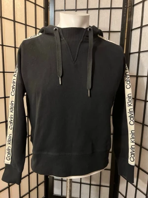 Calvin Klein Performance hoodie – svart, logoband på ärmar (XS) - Svart hoodie från Calvin Klein Performance med logoband längs ärmarna. Enkel modell som passar till det mesta.  Mjuk på insidan och skön att ha på sig, bra som vardagshoodie. 	•	Storlek: XS 	•	Skick: Mycket bra, inga hål eller fläckar 	•	Material: Bomull/polyester  Kan skickas. Säg till om du vill ha fler bilder. 