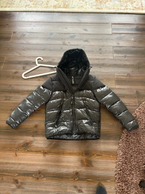 Svart dunjacka från Replay - Snygg svart dunjacka från Replay med glansig finish och huva. Jackan har dragkedja framtill, två sidofickor och quiltad design. Perfekt för kalla dagar och inspirerad av mountain style. Tidlös och trendig look.