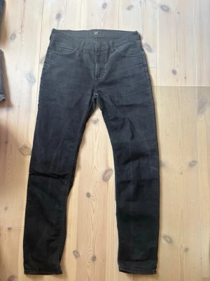 Svarta skinny jeans från Lee - Snygga svarta skinny jeans från Lee med klassisk femficksmodell och hög midja. Jeansen har smal passform och är tillverkade i stretchigt material för extra komfort. Perfekta för en trendig och avslappnad stil. Storlek 33/32