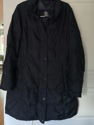 Svart lång dunjacka Tommy Hilfiger - Snygg svart lång dunjacka från Tommy Hilfiger med hög krage och tryckknappar framtill. Jackan har diskret logotyp på ärmen och praktiska fickor. Tvättad och då for dunet ihop så den som orkar ”fluffa” till fodret får en fin jacka för billig peng. 