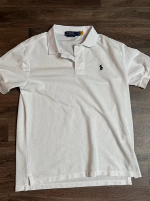 Vit pikétröja från Polo Ralph Lauren - Klassisk vit pikétröja med krage och korta ärmar från Polo Ralph Lauren. Tröjan har den ikoniska broderade loggan på bröstet och är tillverkad i mjuk bomull. Perfekt för en stilren och avslappnad look.