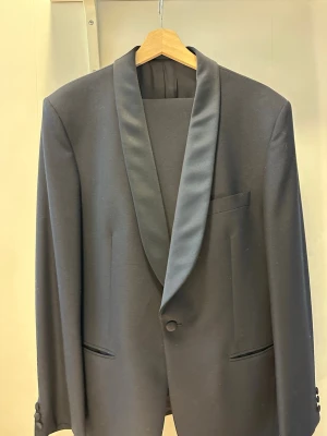 Mörkblå smoking 2 set. - Stilren svart kavaj från Turo med elegant sjalkrage i blankt material. Klassisk design med enkel knappstängning och tre fickor framtill. Detta tillsammans med matchande byxor i samma material och sidenslag längs benet