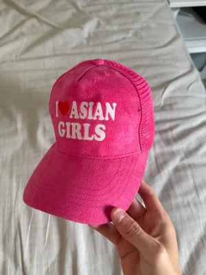 Rosa truckerkeps med text - Säljer en rosa truckerkeps med vit och röd text framtill där det står 'I ♥ ASIAN GIRLS'. Kepsen har böjd skärm, mesh baktill och justerbar snapback. Materialet framtill är mjukt och sammetliknande medan baksidan är i mesh.