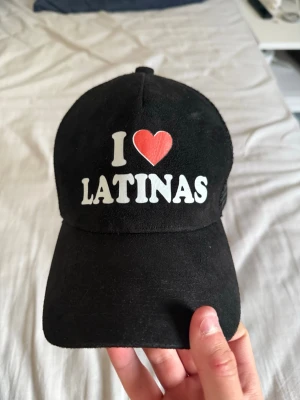 Svart keps med I ❤️ LATINAS-tryck - Säljer en svart keps med vit text och ett rött hjärta där det står 'I ❤️ LATINAS' på framsidan. Kepsen har böjd skärm och justerbar snapback i plast baktill. Materialet är en mix av tyg framtill och mesh baktill för extra ventilation.
