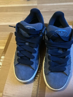 Mörkblå Adidas sneakers i mocka - Snygga mörkblå sneakers från Adidas i mjuk mocka med breda svarta skosnören och klassisk Adidas-logga på plösen. Skorna har en vit sula och en sportig, tidlös design som passar perfekt till streetwear och vardagsstil.