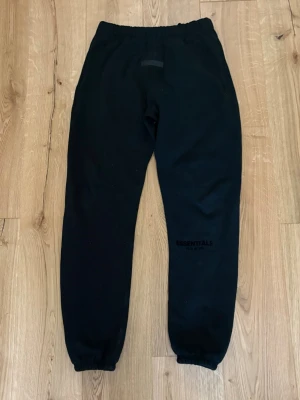 Svarta Essentials mjukisbyxor - Säljer ett par svarta mjukisbyxor från Essentials med resår i midjan och vid bensluten. Byxorna har en enkel och clean design med diskret logga på ena benet. Perfekta för chill dagar eller när du vill ha det bekvämt.