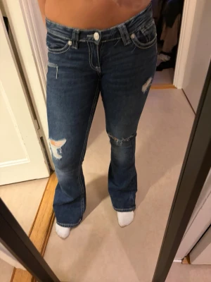 Blå bootcut jeans med slitningar - Säljer ett par blå bootcut jeans med slitningar på låret och knät. Jeansen har vita kontrastsömmar och snygga broderade detaljer på bakfickorna. Klassisk femficksmodell i denim med låg midja och knappgylf.