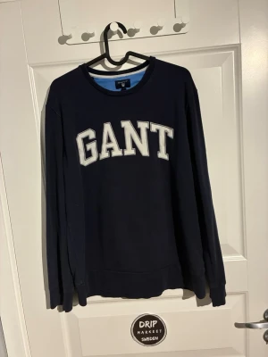 Mörkblå sweatshirt från GANT - Mörkblå sweatshirt från GANT med stora vita bokstäver broderade över bröstet. Tröjan har rund halsringning, långa ärmar och ribbade muddar vid ärmslut och nederkant. Perfekt för en avslappnad och stilren look.