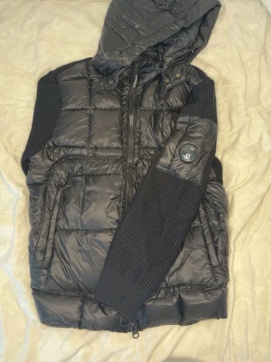 C.P. Company cardigan - Svart cardigan från C.P. Company med vadderad front och huva i glansigt material. Cardiganen har stickade ärmar, dragkedja, fickor och den klassiska linsen på ärmen. Perfekt för en modern streetwear-look.