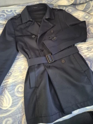 Zara rock  - En Zara mörkblå trenchcoat med klassisk dubbelknäppning, axelklaffar och matchande bälte i midjan (det går att ta bort bältet om man inte vill ha den). Jackan har bred krage och raka ärmar. Materialet ser ut att vara en slät, tålig textil som passar perfekt till en trendig och tidlös look.