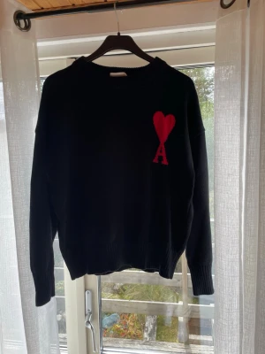 Svart stickad tröja från Ami - Svart stickad tröja från Ami Alexandre Mattiussi med röd hjärtlogga och bokstaven 'A' på bröstet. Tröjan har rund halsringning, ribbade muddar och är tillverkad i mjukt material. Perfekt för dig som gillar stilrena plagg med en twist. Den är i storlek M men råkade tvätta den i för höga grader så skulle säga att den är mer S nu 