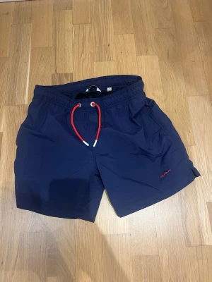 Marinblå badbyxor med röd dragsko - Snygga marinblå badbyxor från Gant med elastisk midja och röd dragsko framtill. Klassisk modell med sidofickor och en bakficka. Tillverkade i snabbtorkande material, perfekta för stranden eller poolen.