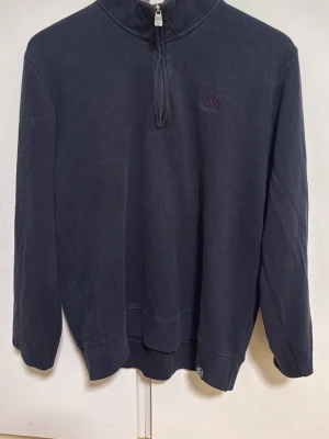 Mörkblå halvzip från Jean Paul - Mörkblå tröja från Jean Paul med halv dragkedja och broderad logga på bröstet. Insidan av kragen är ljusgrå med blått tyg och vinröda detaljer. Perfekt för lager på lager och har ribbade muddar vid ärmslut och nederkant.