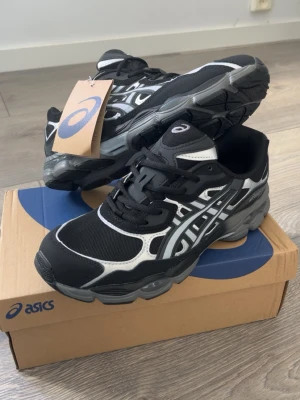 Svarta Asics sneakers med vita detaljer - Snygga svarta Asics sneakers med vita och grå detaljer. Skorna har en sportig design med mesh och syntetmaterial, samt den klassiska Asics-loggan på sidan. Sulan är robust och ger bra stöd. Perfekta för dig som gillar streetwear och sportig stil.