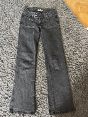 Grå bootcut jeans med dubbla knappar - Snygga grå bootcut jeans med dubbla knappar i midjan och framfickor. Jeansen har en klassisk femficksdesign och utsvängda ben. Perfekta för dig som gillar en retroinspirerad look.