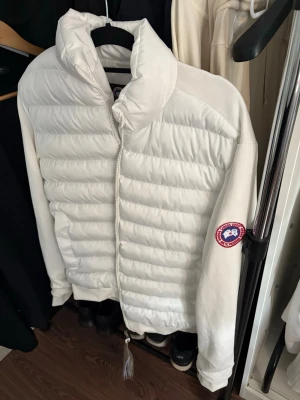 Vit pufferjacka från Canada Goose - Snygg vit pufferjacka från Canada Goose med quiltad design och hög krage. Jackan har dragkedja framtill, två sidofickor och den klassiska Canada Goose-loggan på ärmen. Perfekt för kalla vinterdagar och riktigt stilren look.