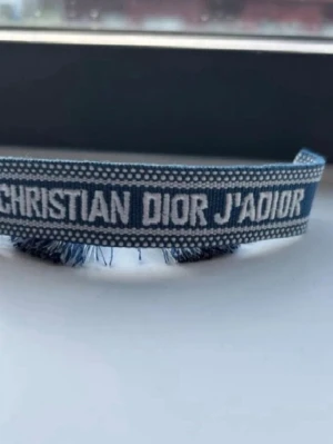 Blått tygarmband Christian Dior - Säljer ett blått tygarmband från Christian Dior med texten 'J'ADIOR' broderad i vitt. Armbandet har fransade kanter och ett stilrent, trendigt utseende. Perfekt accessoar för dig som gillar exklusiva märken och vill sticka ut.