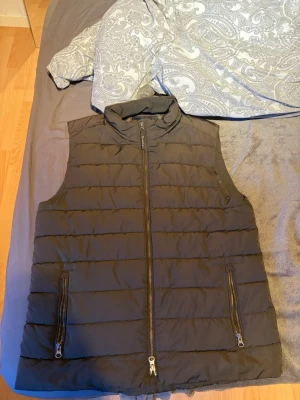 Grön väst bondelid - Snygg grön dunväst med horisontella sömmar och hög krage. Västen har två framfickor med dragkedja och stängs med en kraftig dragkedja framtill. Perfekt att slänga över en hoodie för extra värme. Materialet är quiltat och ger en modern look.