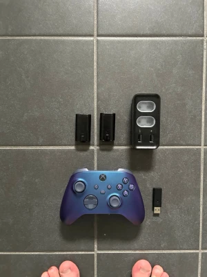 Blå Xbox-handkontroll med tillbehör - Säljer en snygg blå Xbox-handkontroll med matchande laddstation, två uppladdningsbara batteripack och en USB-dongel. Handkontrollen har en modern design med svarta och blå knappar, perfekt för gaming på konsol eller PC.